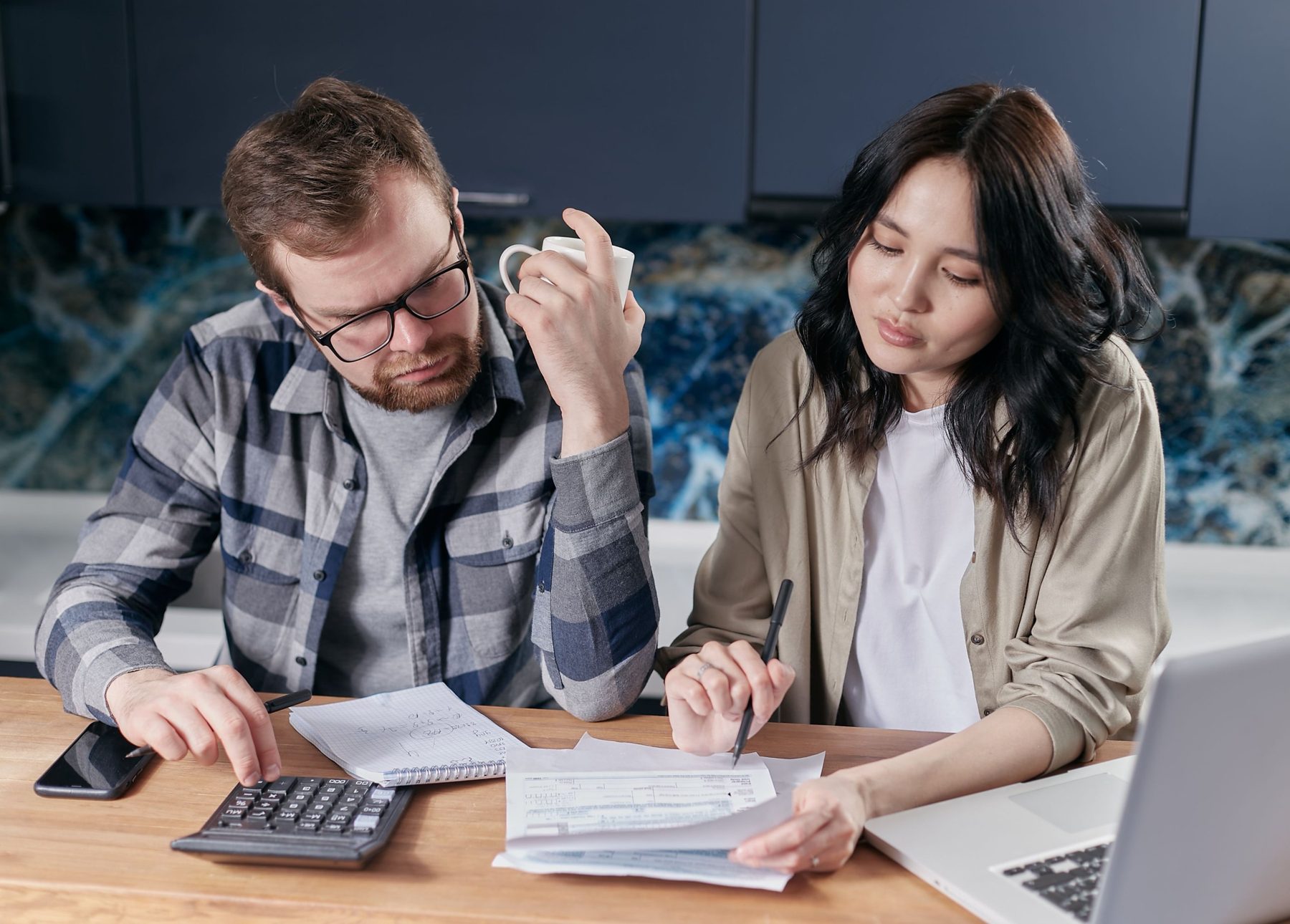 A couple sorting tax forms at a kitchen table with a calculator, notebook and laptop